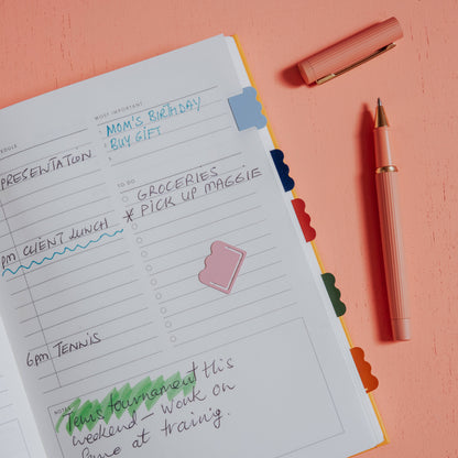 Open planner with notes, pen, Papier Metal Index Tabs (Cloud), on a peach-colored desk.